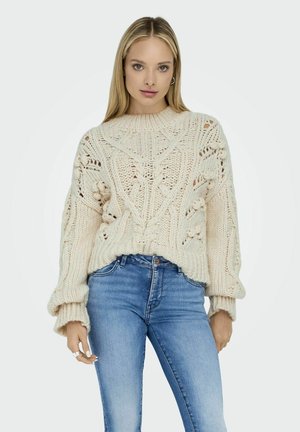 Pull en tricot crème avec des motifs en torsades complexes et des manches surdimensionnées, associé à un jean bleu. Le matériau semble doux et volumineux.