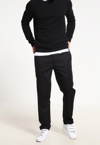 Zwarte trui met lange mouwen over een witte onderhemd, gecombineerd met zwarte broek en witte sneakers. Casual, comfortabele fit.