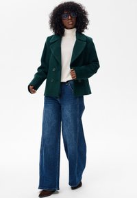 Kobieta z kręconymi włosami, ubrana w oversize'owy ciemnozielony płaszcz, biały golf, szerokie niebieskie dżinsy, ciemne botki i duże okulary przeciwsłoneczne, stoi.