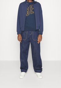 Chaqueta navy con cremallera, sudadera azul con texto amarillo, jeans oscuros de corte holgado y zapatillas blancas. Diseño simple con detalles en contraste.