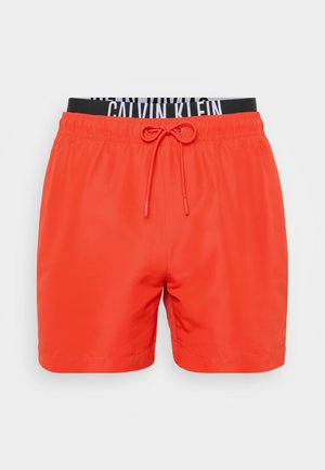Sarkani peldkostīmi, kas izgatavoti no viegla materiāla, ar elastīgu jostasvietu, kurā ir melnbaltā Calvin Klein logo un aukliņu detaļa.