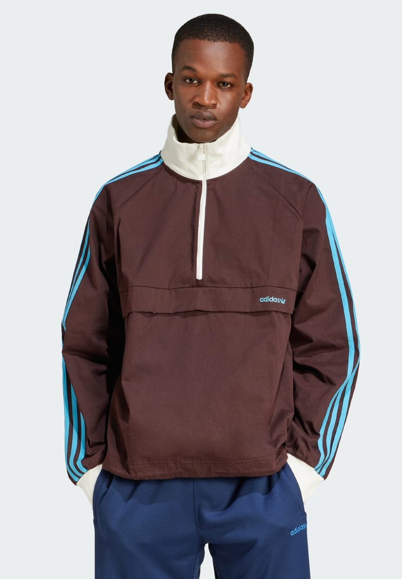 Pullover castanho com zíper até à metade, com colarinho branco e riscas azuis nas mangas. Apresenta o logótipo da Adidas na frente e um bolso tipo pouch.