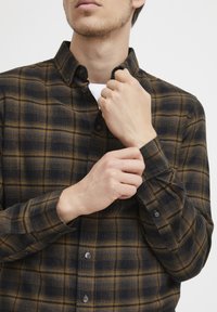 Camisa de botones con un patrón de cuadros en colores azul oscuro y marrón. La tela parece suave. Cuenta con un cuello y mangas con puños abotonados.