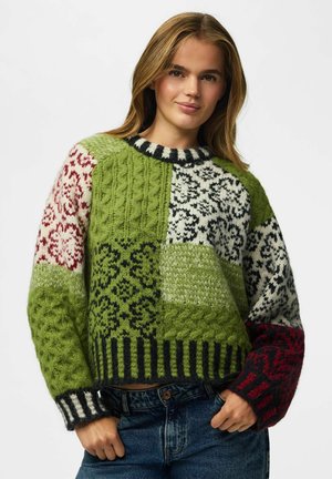 Jeune femme aux longs cheveux blonds portant un pull en tricot multicolore vert, noir, blanc et rouge à motifs, et un jean bleu.