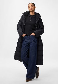 Donna che indossa un lungo piumino nero, camicia nera con bottoni, jeans a gamba larga blu e scarpe marrone scuro, in piedi con le mani nelle tasche.