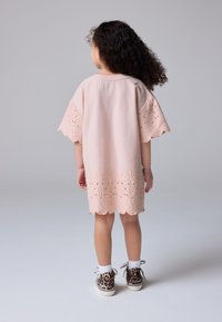 Svetloružové šaty s perforovanými rukávmi a zaobleným lemom, zdobené prepracovanou výšivkou. Kombinované s teniskami so vzorom leopardieho kožušiny.