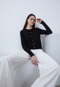 Cardigan nero con collo rotondo, chiusura a bottoni e maniche lunghe. Indossato sopra pantaloni bianchi a gamba larga, su uno sfondo chiaro.