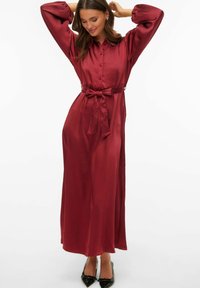 Długa bordowa sukienka z satyny z kołnierzykiem, zapinana na guziki z przodu, z elastycznymi bufkami na rękawach i wiązanym paskiem w talii. Noszona z czarnymi butami na obcasie o ostrym nosku.