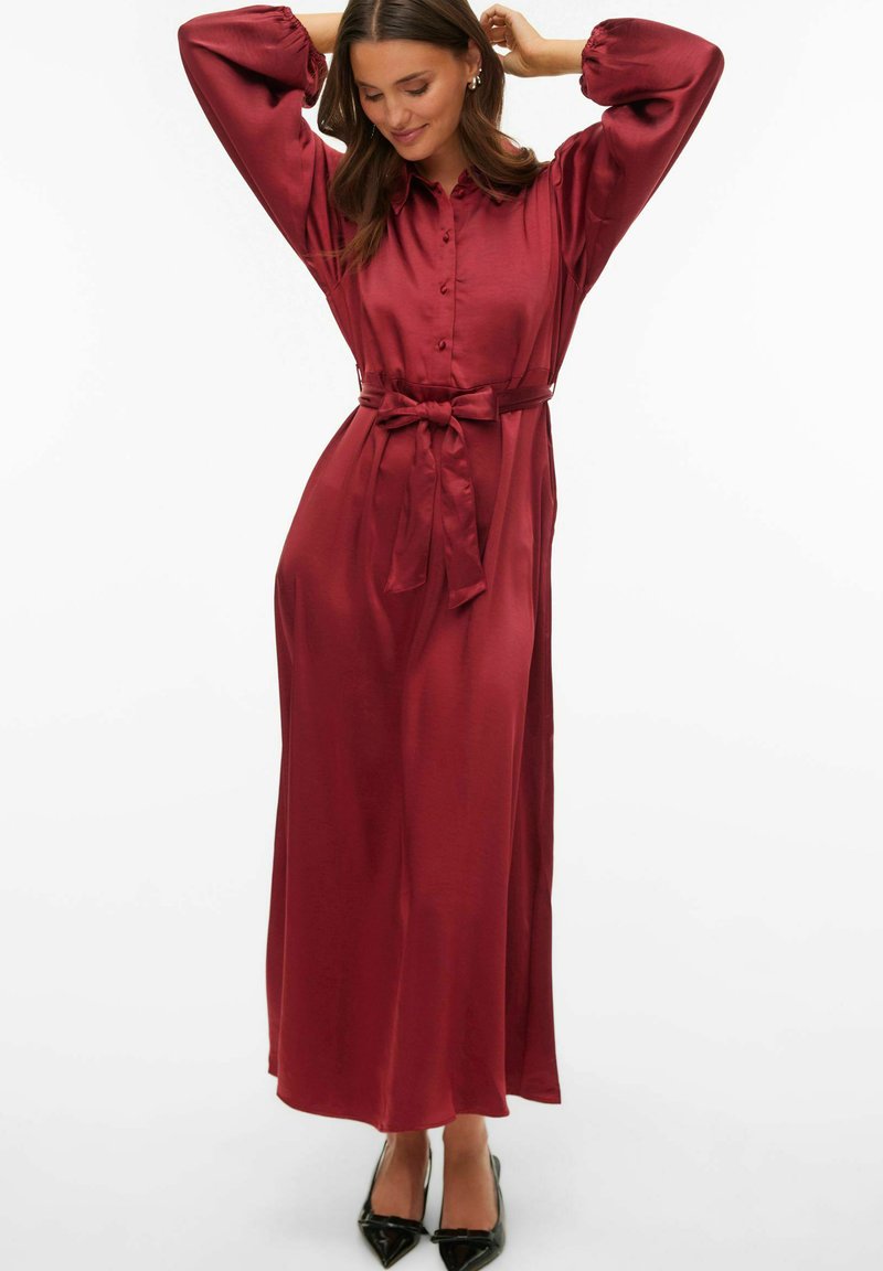 Długa bordowa sukienka z satyny z kołnierzykiem, zapinana na guziki z przodu, z elastycznymi bufkami na rękawach i wiązanym paskiem w talii. Noszona z czarnymi butami na obcasie o ostrym nosku.