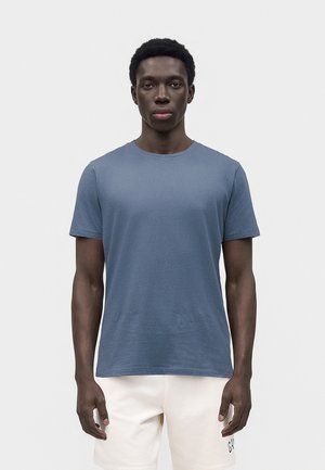 Νεαρός άνδρας που στέκεται με το πρόσωπο προς τα εμπρός φορώντας μπλε κοντομάνικο μπλουζάκι και ανοιχτόχρωμα σορτς σε απλό φόντο.