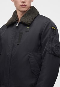 Homme portant une veste bomber noire avec un col en polaire vert foncé, une poche zippée sur la manche, des poches latérales et de petits écussons logo jaunes sur la poitrine et le bras.