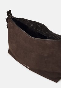 Hnědá semišová kabelka se zipovým uzávěrem, která má vnitřní prošívaný vzor. Design zahrnuje ploché dno a diskrétní detaily loga.