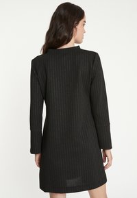 Schwarzes Kleid mit langen Ärmeln, ausgestattet mit einem silbernen Streifenmuster. Gerader Schnitt mit hohem Hals und sichtbaren Nahtdetails.