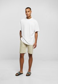 Camiseta blanca de algodón, corte relajado. Pantalones cortos color bronceado, de longitud media. Sandalias negras con texto blanco. Textura suave, diseño minimalista y colores neutros.