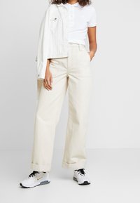 Pantalons larges beige clair avec des ourlets retroussés, associés à un polo blanc et une veste en denim blanche. En ce qui concerne les chaussures, il s'agit de baskets blanches.