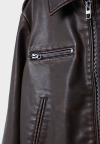 Ciemnobrązowa kurtka skórzana z kieszenią na piersi zapinaną na srebrny zamek błyskawiczny oraz pionową kieszenią z przeszyciami poniżej, wyposażona w duże zapięcie na zamek błyskawiczny z przodu.