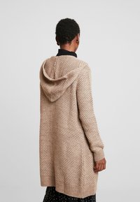 Beżowy dziergany kardigan z fakturowanym wzorem, długimi rękawami i kapturem. Sięga do połowy uda, posiada ściągacze na mankietach i dolnym brzegu.