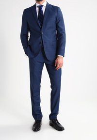 Abito blu navy realizzato in tessuto texturizzato, con giacca a due bottoni e pantaloni slim-fit. Abbinato a una camicia bianca e una cravatta a righe blu.