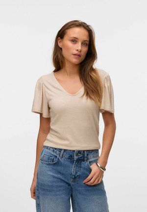 T-shirt beige à col en V avec des manches courtes bouffantes. Texture douce, coupe ajustée, associé à un jean bleu taille haute et un bracelet en argent au poignet.