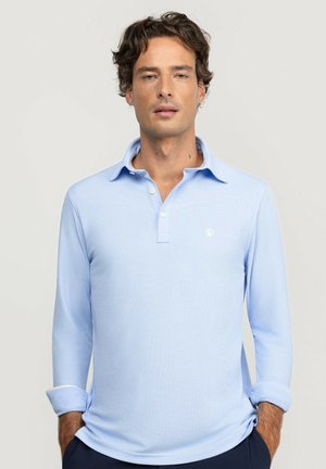 Hombre con cabello rizado que lleva una camisa polo de manga larga azul claro con cuello y botones, manos en los bolsillos de pantalones oscuros, fondo neutro.