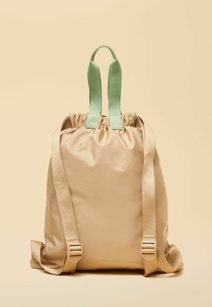 Beiger Rucksack mit Kordelzug, breiten mintgrünen Tragegriffen und verstellbaren beigen Schultergurten vor hellem Hintergrund.