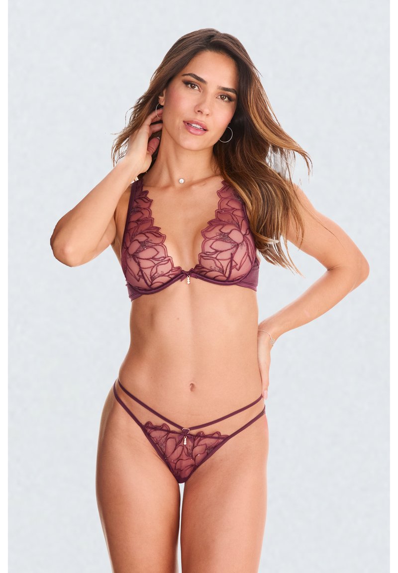 Woman wearing sheer burgundy lace lingerie set with floral embroidery, standing with one hand on neck and the other on hip against plain background.