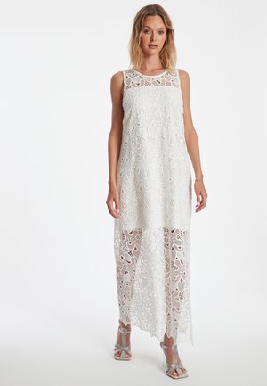 Robe maxi blanche en dentelle avec des motifs floraux, sans manches, avec un ourlet transparent et des détails raffinés. Associée à des talons en argent scintillants.