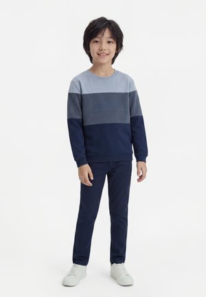 Dreng står og smiler iført en blåt farveblok-sweatshirt, marineblå bukser og hvide sneakers på en hvid baggrund.
