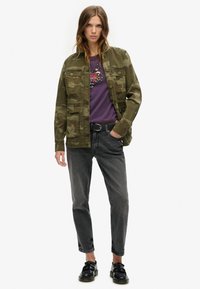 Chaqueta de camuflaje verde oliva con cuatro bolsillos, llevada encima de una camiseta gráfica púrpura. Combinada con jeans grises y zapatos negros de cordones.