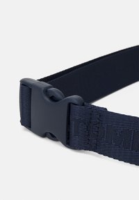 Marinblå nylonbälte med plastspänne. Har en vävd textur och ett präglat logotyp på remmen. Idealisk för säker fastsättning.