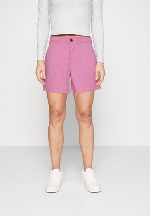 Femme portant un crop top blanc à manches longues, un short rose avec poches avant, et des baskets blanches, debout devant un fond clair uni.