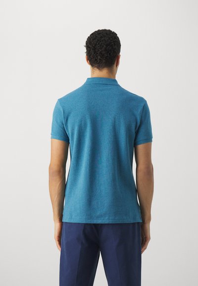 Camisa polo turquesa de tejido texturizado, con cuello clásico y mangas cortas, combinada con pantalones azul marino. Se muestra vista trasera.