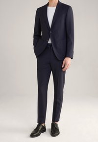Granatowy garnitur z dopasowaną marynarką i szytymi spodniami, w połączeniu z białą koszulką i czarnymi eleganckimi butami. Gładki materiał z minimalnymi detalami.