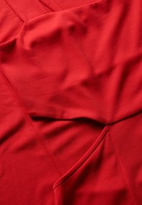 Czerwona tkanina o gładkiej teksturze, z drapowanymi fałdami i detalem kieszeni. Miękki, elastyczny materiał w jednolitym kolorze i z czystymi liniami.
