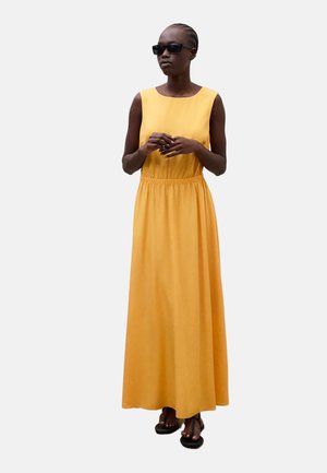 Frau mit geflochtenem Haar, die schwarze Sonnenbrille, ein ärmelloses gelbes Maxikleid und schwarze Sandalen trägt, stehend mit gefalteten Händen.