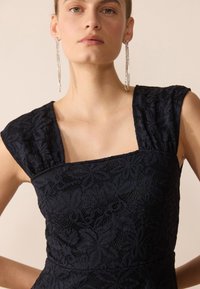 Laivastonsininen pitsimekko, jossa on neliön muotoinen pääntie, lyhyet hihat ja kukkakuviot. Korostettu pitkillä, hopeisilla korvakoruilla lisätyn eleganssin vuoksi.