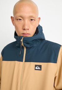 Close-up of a bald person wearing a tan and navy hooded jacket with a front zipper and a logo patch on the chest.