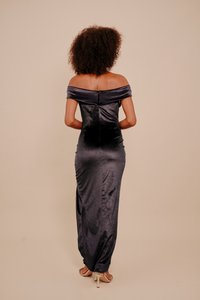 Göndör hajú nő hosszú, vállvillantós sötétlila szatén ruhában és fehér magassarkú szandálban, egyszínű háttér előtt állva.