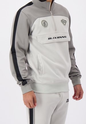 Hombre vistiendo un chándal gris y blanco con rayas negras, parches y el texto "BLCK BNNS" en la chaqueta y los pantalones.