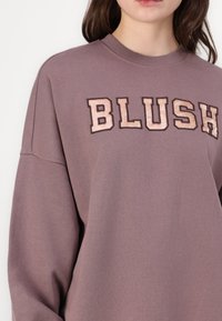 Prevelik, ružičast sivi dukserica od meke mješavine pamuka. Sadrži "BLUSH" u teksturiranom bež pismu. Ribljenje oko vrata i dizajn rukava.
