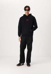 Melns kapučins ar sarkanu "Santa Cruz" logo, kangaroo kabata, melnas bikses un melni apavi ar gumijas zoles. Modelis valkā saulesbrilles.