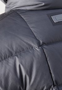 Gros plan sur un tissu matelassé gris foncé avec des coutures visibles et une texture subtile, montrant une petite pièce rectangulaire cousue dessus.