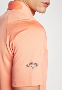 Kortermet oransje polo skjorte med teksturert stoff. Har Callaway-logo brodert på venstre erme og en foldet krage.