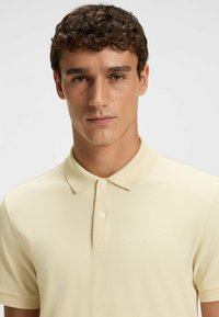 Ung man med kort lockigt hår som bär en ljusbeige pikétröja med två knappar och en subtil "BOSS"-logga på bröstet.