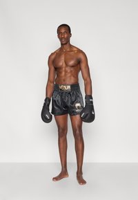 Juodi satino bokso šortai su auksiniu „Venum“ logotipu, juodos bokso pirštinės ir vyriškas modelis be marškinėlių, stovintis basas prieš neutralią foną.