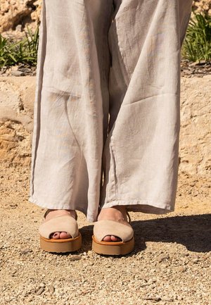Nohy v béžových sandáloch s otvorenými špičkami a hnedými podrážkami, stojace na štrku vedľa kamennej steny, béžové nohavice s širokými nohavicami zakrývajúce členky.