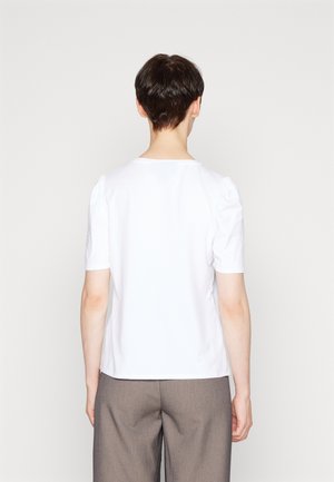 Personne aux cheveux courts et foncés portant un haut blanc à manches courtes uni et un pantalon gris, debout contre un fond neutre clair, vue de dos.