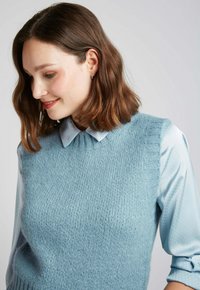 Gilet en tricot texturé bleu clair avec un col côtelé, porté par-dessus une chemise à pois bleu clair. Tissu doux, design décontracté, sans motifs visibles.