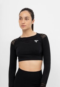 Sort langærmet cropped top med tekstureret stof, mesh-detaljer på skuldrene og et hvidt logo på venstre side. Mat finish.