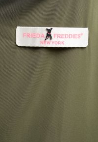 Biała metka z różowym napisem "Frieda & Freddies New York" oraz małym czarnym psem na zielonym tle.
